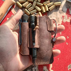 May include: A brown leather EDC organizer with a flashlight, pen, and a blue clip. The organizer is held in a hand, with a knife and bullets in the background. The leather has a distressed look.