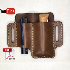 Może przedstawiać: Brązowa skórzana pochwa z dwoma kieszeniami, jedna z czarno-niebieskim długopisem, a druga z drewnianym narzędziem. Pochwa ma pętlę na pasek i jest zszyta białą nicią.