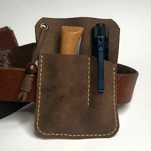 May include: A brown leather belt pouch with a pocket containing a wooden knife handle and a blue flashlight. The pouch is stitched with light-colored thread and has a leather strap with a wooden bead.