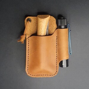 Könnte beinhalten: Ein braunes Ledertasche mit einem kleinen Messer und einer Taschenlampe im Inneren. Die Tasche hat eine Schlaufe zum Befestigen an einem Gürtel.