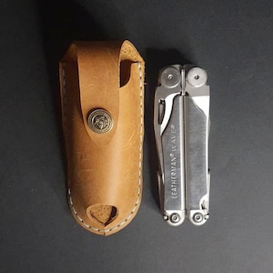 May include: A silver Leatherman Wave multi-tool with a brown leather sheath. The multi-tool is open and the pliers are extended. The sheath has a decorative button with a star design.