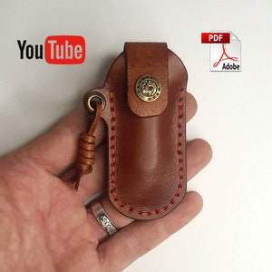 May include: Brown leather knife sheath with a red stitching detail and a brass snap closure. The sheath has a loop for attaching a key ring. The sheath is being held in a hand.
