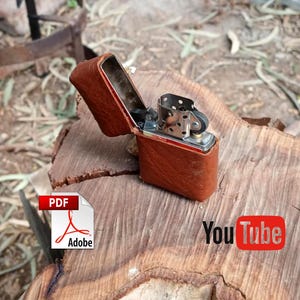 May include: A brown leather lighter case with a silver lighter inside. The case is open and the lighter is visible. The lighter has a silver finish and a black wick. The case and lighter are sitting on a wooden surface. The words "You Tube" are visible in the lower right corner of the image.