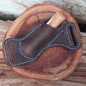 Könnte beinhalten: Eine braune Ledertasche für ein Messer mit blauen Nähten. Die Tasche hat eine Gürtelschlaufe und eine Klappe, die mit einem Druckknopf befestigt wird. Die Tasche ist für ein kleines Messer mit einem Holzgriff konzipiert.