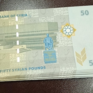 May include: A stack of fifty Syrian pound banknotes. The notes are predominantly light green and white, displaying the Central Bank of Syria logo, a building, and a statue. The number "50" is clearly visible on each note.