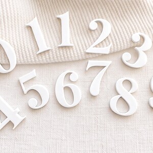 White acrylic number set for monthly baby milestone sign (0–9 plus extra 1), shown on neutral textured fabric background.