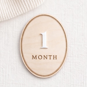 Oval monthly baby milestone sign in natural birch wood with white acrylic “1” and engraved “MONTH,” clean neutral backdrop.