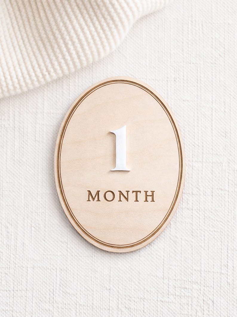 Oval monthly baby milestone sign in natural birch wood with white acrylic “1” and engraved “MONTH,” clean neutral backdrop.
