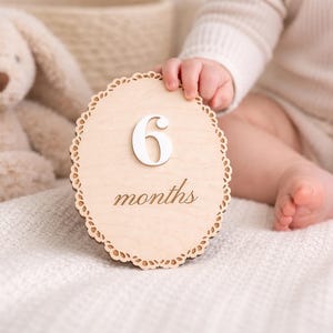 Monthly baby milestone sign photo prop in use, baby holding scalloped birch wood plaque with white acrylic “6” and engraved “months” on neutral bedding.