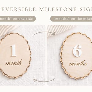 May include: Reversible milestone sign with the text "month" on one side and "months" on the other. The sign is made of light wood and features a decorative scalloped edge. One side shows the number 1, the other shows the number 6.