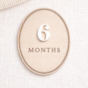 Oval monthly baby milestone sign in natural birch wood with white acrylic “6” and engraved “MONTHS,” photographed on neutral textured fabric.