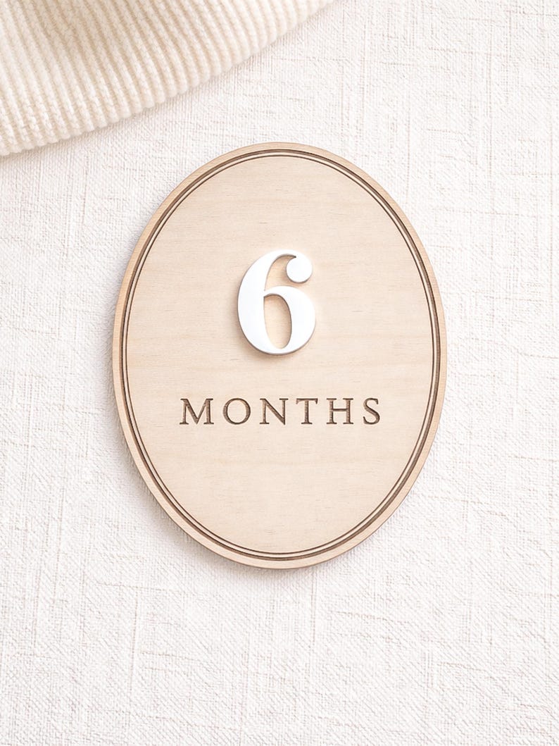Oval monthly baby milestone sign in natural birch wood with white acrylic “6” and engraved “MONTHS,” photographed on neutral textured fabric.
