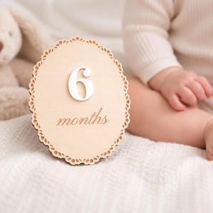 Monthly baby milestone sign used as a newborn photo prop, scalloped birch wood plaque showing “6 months” beside baby on soft neutral blanket.