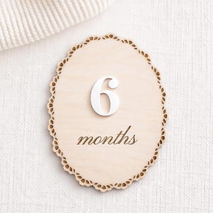 Scalloped monthly baby milestone sign in natural birch wood with white acrylic “6” and engraved script “months,” shown on neutral fabric.