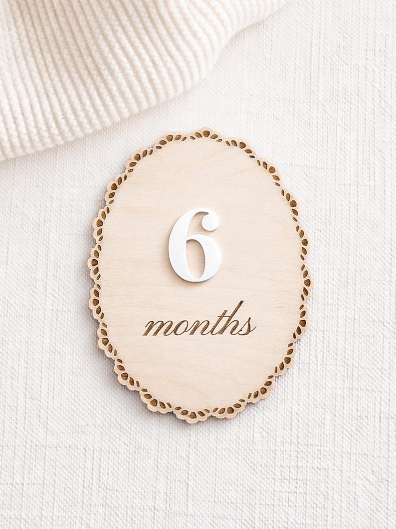 Scalloped monthly baby milestone sign in natural birch wood with white acrylic “6” and engraved script “months,” shown on neutral fabric.