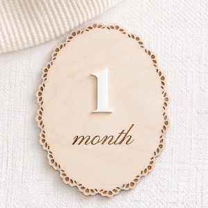 Scalloped monthly baby milestone sign in natural birch wood with white acrylic “1” and engraved script “month,” neutral fabric background.