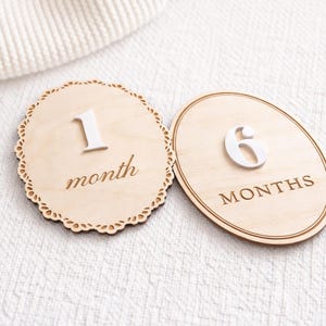Monthly baby milestone sign set showing two styles at an angle—scalloped “1 month” and oval “6 months”—natural birch wood with white acrylic numbers.