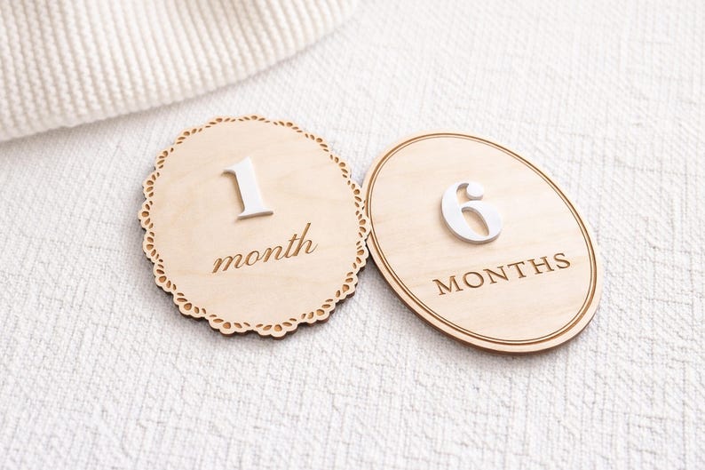 Monthly baby milestone sign set showing two styles at an angle—scalloped “1 month” and oval “6 months”—natural birch wood with white acrylic numbers.