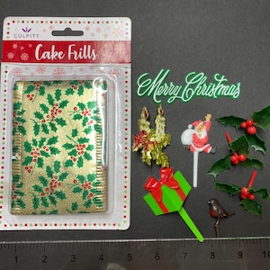 May include: A collection of Christmas cake decorations. Includes a package of gold cake frills with a holly pattern, a "Merry Christmas" sign, Santa Claus, holly sprigs, a gift, candles, and a bird.
