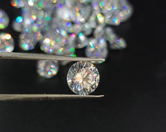 May include: A clear, round, faceted diamond gemstone held by tweezers against a black background. The diamond is surrounded by a scattering of other diamonds.