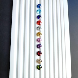 May include: A row of round, faceted gemstones in various colours, including blue, turquoise, purple, red, white, yellow, pink, green, and brown, displayed on a white background.