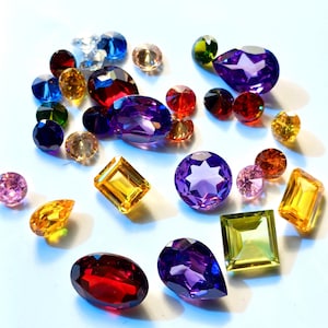 May include: An assortment of colourful gemstones in various shapes and sizes, including purple, red, yellow, and blue. The gemstones are arranged on a white surface.