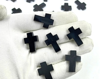 May include: A hand holding a collection of black obsidian cross charms. The crosses are smooth and polished, with a slightly rounded shape.