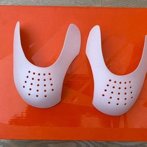 Puede incluir: Dos protectores de pliegues de zapatos blancos con un patr&oacute;n de puntos. Los protectores est&aacute;n sobre una superficie naranja brillante.