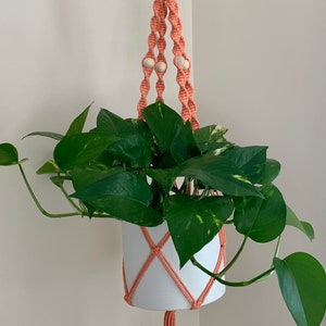 Op de afbeelding: Een perzikkleurige macramé plantenhanger met houten kralen en een witte pot met een groene plant erin.