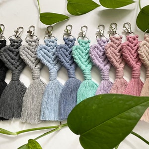 May include: A set of ten colourful macrame heart keychains with tassels. The keychains are made with a variety of colours, including black, grey, light blue, blue, green, purple, pink, and beige.