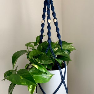 Peut inclure: Un porte-plante en macramé bleu marine avec des perles en bois et un pot blanc avec une plante verte.