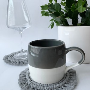 Op de afbeelding: Twee grijze gehaakte onderzetters met franjes, een met een grijs en wit mok erop en de andere met een helder wijnglas erop.