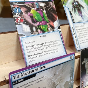 May include: A purple and green superhero card game featuring Kang the Conqueror. The card shows Kang in a purple and green costume with a large, menacing expression. The card text reads "The Master of Time" and includes the text "Forced Interrupt" and "When Defeated".
