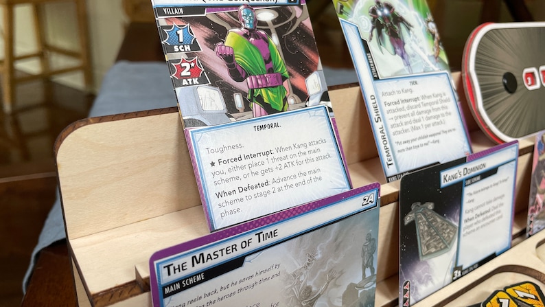 May include: A purple and green superhero card game featuring Kang the Conqueror. The card shows Kang in a purple and green costume with a large, menacing expression. The card text reads "The Master of Time" and includes the text "Forced Interrupt" and "When Defeated".
