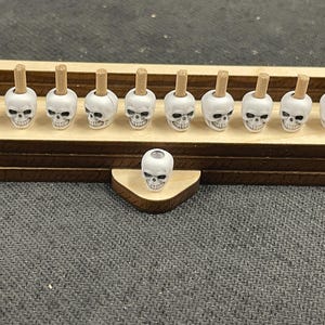 May include: A wooden display piece with a dark brown base and a light brown top, holding eleven small white skull-shaped objects with wooden stems. A single skull sits in front. The skulls have black eye sockets and noses.