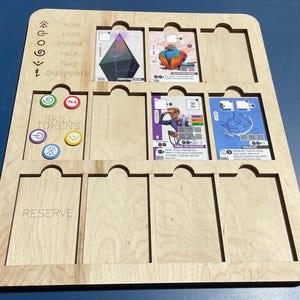 May include: A wooden game board with slots for cards and skill tokens. The board has text that reads "MOVE, LOOK, ENGAGE, HELP, TAKE, OVERPOWER" and "RESERVE". Several game cards and tokens are visible.
