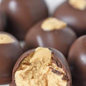 May include: Close-up of chocolate-covered peanut butter balls. The dark chocolate coating is smooth and glossy, with a peanut butter filling visible in one partially bitten treat. The peanut butter filling is light brown and textured.
