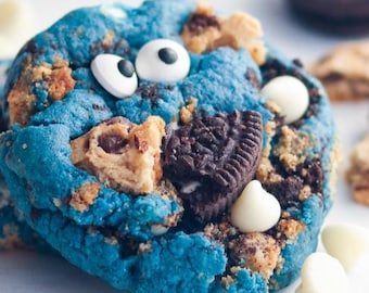 May include: A close-up of a blue cookie with white candy eyes, chocolate chip pieces, and an Oreo cookie. White chocolate chips and cookie crumbs are scattered around the cookie. The cookie has a textured appearance.