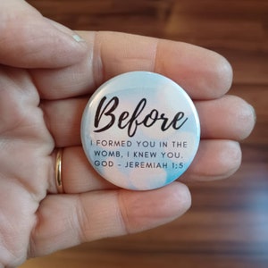 May include: A white button with a blue and pink watercolor background. The button has black text that reads "Before I formed you in the womb, I knew you. God - Jeremiah 1:5"