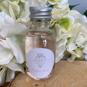 May include: A clear glass bottle with a silver lid and a white label that says "WB Weave & Bloom English Rose". The bottle is sitting on a wooden surface with white flowers in the background.