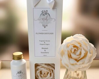 May include: A white rectangular box with a satin bow, labelled Flower Diffuser, Rhubarb & Strawberry, 100 ml. Beside it sits a white bottle of diffuser oil and a clear glass vase containing a large, cream-coloured artificial rose flower diffuser.