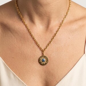 May include: A gold-toned chain necklace featuring a circular pendant with a sunburst design and a central blue and white evil eye motif. The pendant hangs from a textured link chain that rests against the collarbone area of the neck.