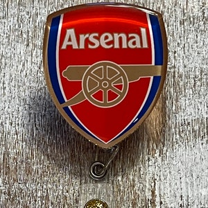 May include: A red, white, and gold Arsenal logo badge reel. The badge features the Arsenal cannon logo and the word "Arsenal" in white. The reel has a gold glitter accent and a metal clip.
