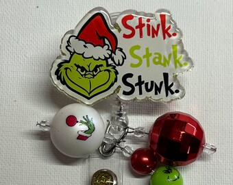 May include: A Grinch-themed badge reel with a clear, glitter-edged top featuring the Grinch's face and the words "Stink. Stank. Stunk." in red and green. Includes decorative beads and a red disco ball.