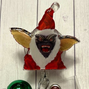 May include: A badge reel featuring a Gremlin character in a Santa Claus outfit. The design includes a red Santa hat, a white beard, and a menacing expression. The badge reel has a clear plastic covering and a metal clip.