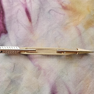 May include: A gold-colored tie clip in the shape of an arrow. The arrow has a detailed fletching design on one end and a pointed tip on the other. The clip is set against a colorful, abstract background.