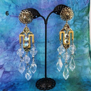 May include: Ornate gold-toned earrings with clear crystal accents. Each earring features a circular top, a decorative center, and dangling crystal beads. The earrings are displayed on a black stand against a blue background.