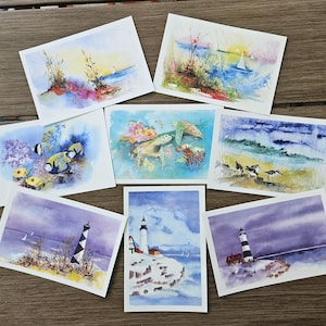 Peut inclure: Plusieurs peintures à l'aquarelle sur papier blanc. Les peintures représentent diverses scènes côtières, notamment des phares, la vie marine et des voiliers. L'œuvre présente des bleus, des verts et des violets, capturant l'essence de l'océan.