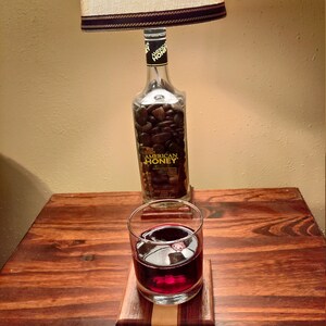 May include: A table lamp with a beige lampshade and a dark brown trim. The lamp's base is a repurposed Wild Turkey American Honey bottle filled with dark stones. A glass of red liquid sits on a wooden coaster.