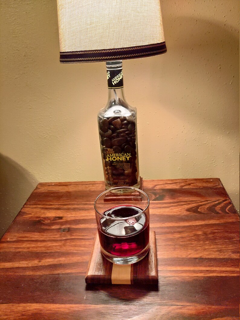 May include: A table lamp with a beige lampshade and a dark brown trim. The lamp's base is a repurposed Wild Turkey American Honey bottle filled with dark stones. A glass of red liquid sits on a wooden coaster.